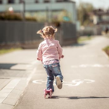 Two years old girl riding her scooter on the street. back view