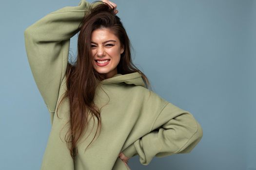 Young beautiful woman looking at camera. Trendy girl in casual hipster hoodie clothes. Positive female shows facial emotions. Funny model isolated on blue background with free space for text.
