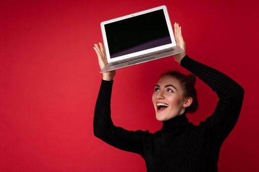 Photo of Beautiful smiling happy young brunette woman holding computer laptop with empty monitor screen wearing black longsleeve looking up at netbook isolated over red background. Mock up