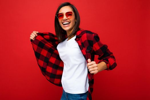 Portrait of positive cheerful smiling young brunette woman in casual white t-shirt for mockup, stylish red check shirt and trendy red sunglasses isolated on red background with copy space.