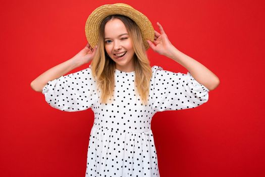 Photo shot of young beautiful cute happy blonde woman wearing casual dress and straw hat isolated over red background with copy space.