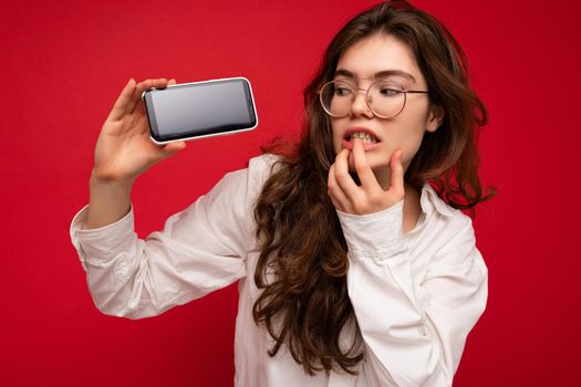 Thoughtful attractive young brunet woman wearing white shirt and optical glasses isolated over red background holding in hand and showing mobile phone with empty screen for mockup looking at gadjet screen and being concerned.