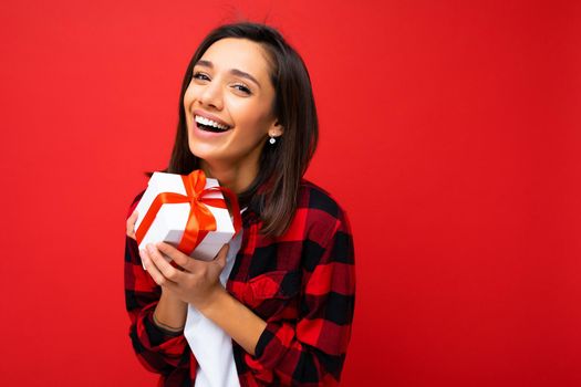 Shot of attractive positive smiling young brunette woman isolated over colourful background wall wearing everyday trendy outfit holding gift box and looking at camera.