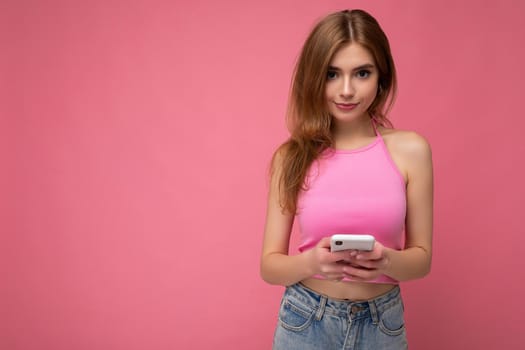 Photo of good looking young blonde woman wearing pink top poising isolated on pink background with empty space holding in hand and using mobile phone messaging sms looking at camera. copy space