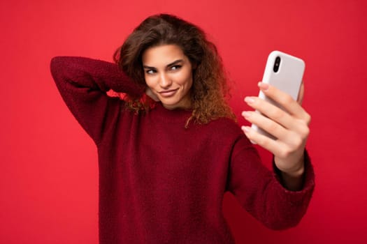 Beautiful young woman with curly hair wearing dark red sweater isolated on red background wall holding and using smart phone looking at telephone screen and taking selfie.