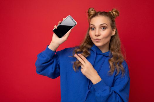 Attractive smiling young blonde woman wearing stylish blue hoodie isolated on red background with copy space holding smartphone showing phone in hand with empty screen display looking at camera showing.