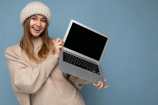 Close-up photo portrait of beautiful smiling young blond woman in winter warm knitted beige hat holding computer laptop looking at camera wearing beige winter sweater isolated over light blue wall background in studio.