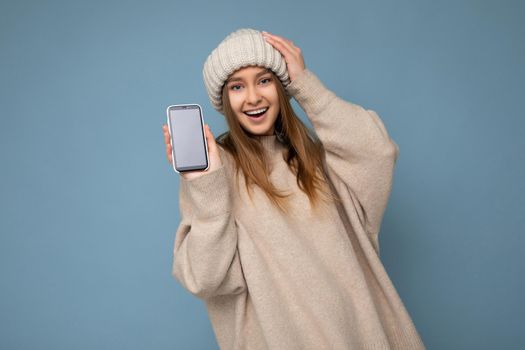 Photo of beautiful smiling young woman good looking wearing casual stylish outfit standing isolated on background with copy space holding smartphone showing phone in hand with empty screen display for mockup looking at camera.