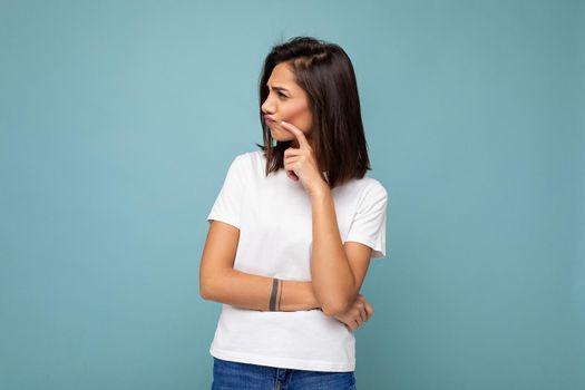 Portrait of thoughtful young pretty nice winsome brunette woman with sincere emotions wearing casual white t-shirt for mockup isolated over blue background with copy space.