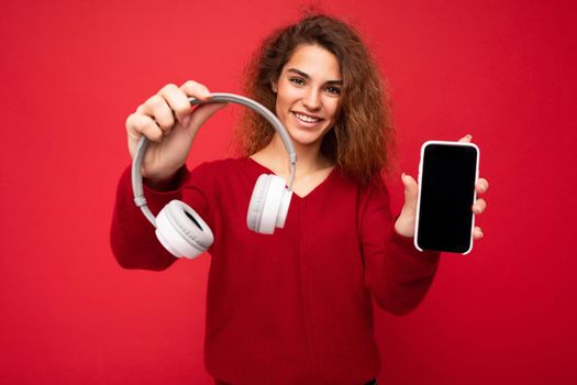 Photo of beautiful positive smiling young woman wearing stylish casual outfit isolated on colorful background wall holding white bluetooth wireless earphones and showing mobile phone with empty screen for mockup looking at camera.