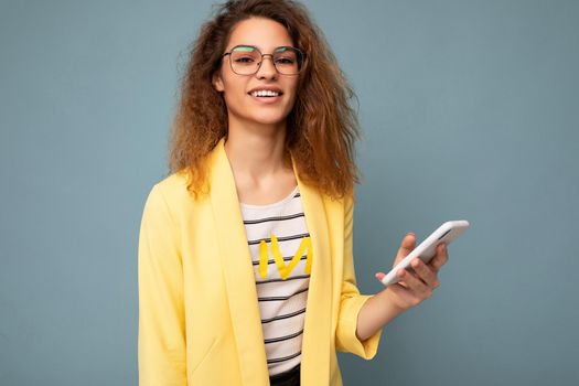 Portrait of attractive young woman with curly dark blond hair wearing yellow jacket and optical glasses isolated on background holding and using phone looking at camera.