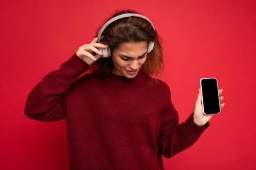 Beautiful happy smiling young woman wearing stylish casual outfit isolated on background wall holding and showing mobile phone with empty display for mockup wearing white bluetooth headphones listening to music and having fun looking down.