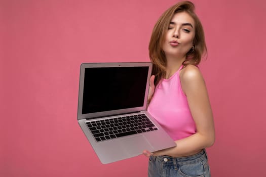 Close-up photo of Beautiful young blond woman holding computer laptop wearing pink crop top looking at camera with close eyes and giving a kiss isolated on pink background. Mock up, copy space, cutout