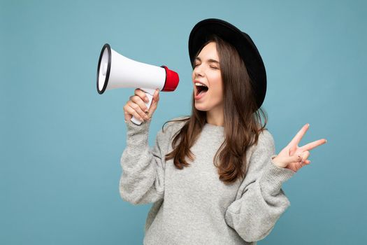 Photo of young beautiful positive brunette woman with sincere emotions wearing stylish black hat and grey sweater isolated over blue background with copy space and screaming in magaphone, shows peace gesture.