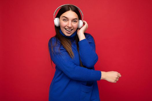 Attractive positive young brunette woman wearing blue sweater isolated over red background wall wearing white bluetooth earphones listening to cool music and having fun looking at camera.