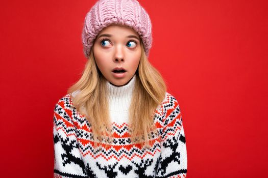 Photo of beautiful shocked young blonde woman isolated over red background wall wearing winter sweater and pink hat looking to the side.