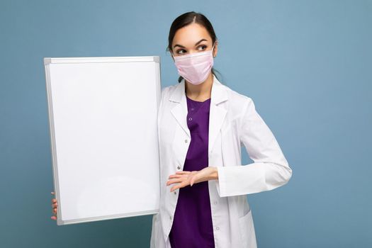 Beautiful brunette female nurse in protective face mask and white medical coat holding an empty magnetic board isolated on blue background.