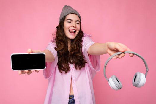 Photo of beautiful positive smiling young woman wearing stylish casual outfit isolated on colorful background wall holding white bluetooth wireless earphones and showing mobile phone with empty screen for mockup looking at camera.