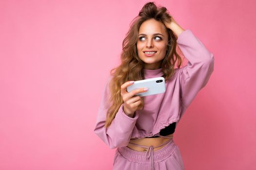 Sexy attractive blonde young woman wearing pink hoodie isolated over pink background with copy space holding and using mobile phone looking to the side.