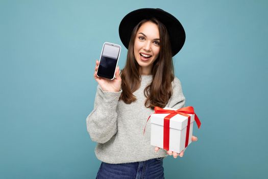 Shot of pretty smiling positive young brunette woman isolated over blue background wall wearing stylish black hat and grey sweater holding gift box showing smartphone screen display for mockup and looking at camera.