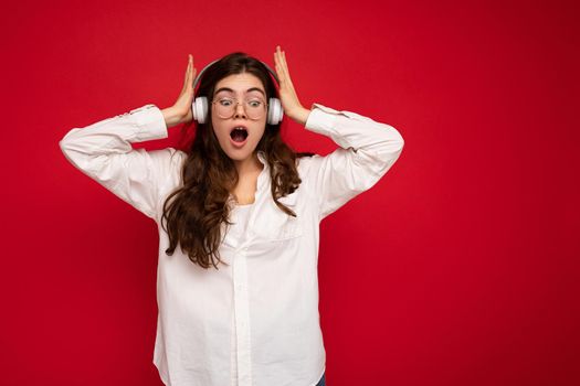 Attractive shocked young brunette female person wearing white shirt and optical glasses isolated over red background wearing white wireless bluetooth earphones listening to music and looking down to the side with open mouth.