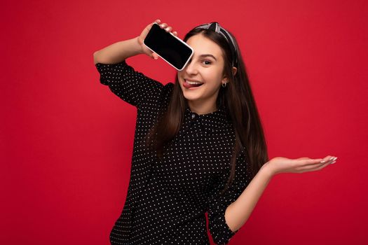 Delightful smiling Pretty brunette girl standing isolated over red wall wearing casual stylish black clothes showing mobile phone with empty screen for cutout looking at camera.