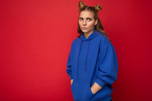 Portrait of young sad upset attractive winsome blonde woman with two horns with sincere emotions wearing casual bright blue hoodie isolated on red background with free space.