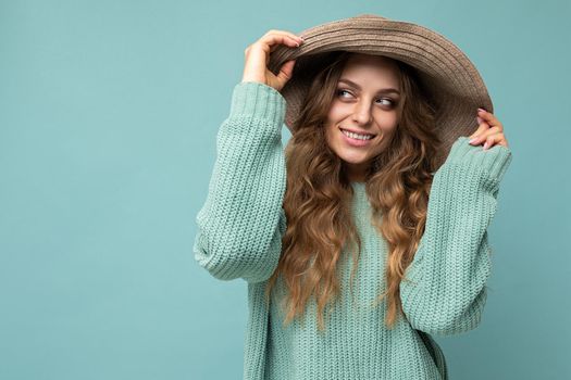 Young beautiful cute curly blonde woman with sexy expression, cheerful and happy face wearing trendy blue sweater and hat isolated over blue background with copy space.