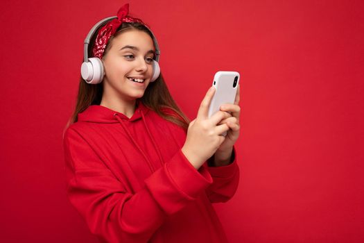 Closeup portrait photo of beautiful happy smiling brunette girl wearing red hoodie isolated on red background holding and using smartphone communicating online wearing white wireless headphones listening to cool music looking at gadjet display.