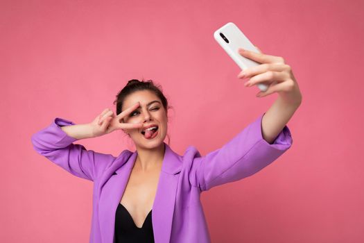 Smiling sexy woman wearing purple suit taking selfie photo on the mobile showing tongue and peace gesture phone isolated over pink background looking at cell display.