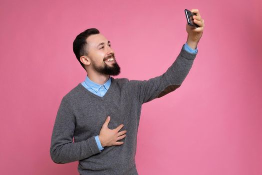 Handsome young man wearing casual stylish clothes standing isolated over background wall holding smartphone taking selfie photo looking at mobile phone screen display.