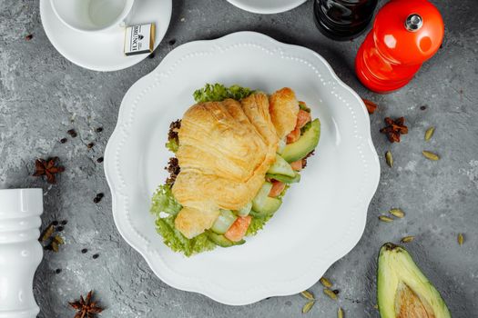 Croissant sandwich with red fish, avocado, fresh vegetables and arugula on black shale board over black stone background. Healthy food concept.