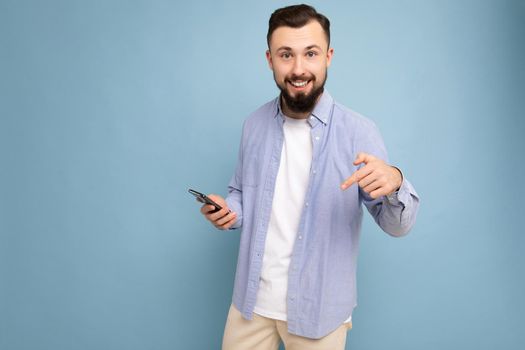 Portrait of happy smiling handsome young brunette unshaven man with beard wearing stylish white t-shirt and blue shirt isolated over blue background with empty space holding in hand and using phone messaging sms looking at camera.