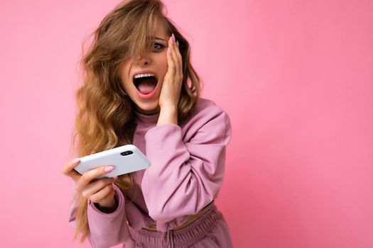 Photo of attractive crazy amazed surprised young woman wearing casual stylish clothes standing isolated over background with copy space holding and using mobile phone looking at camera and shouting.