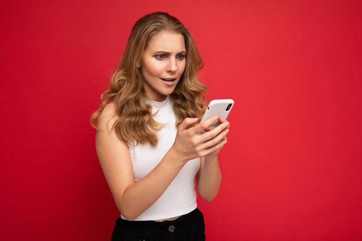 Amazed shocked beautiful young blonde woman wearing white t-shirt isolated over red background using smartphone and chatting via mobile phone looking at gadjet screen.