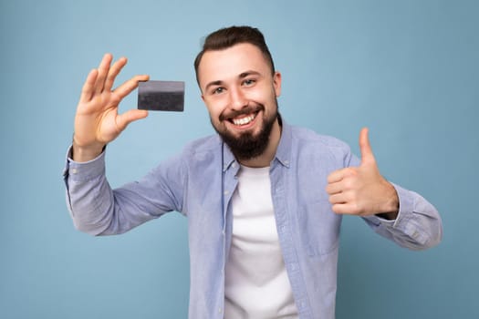 Closeup portrait of Good looking cool smiling brunette bearded young man wearing stylish blue shirt and white t-shirt isolated over blue background wall holding credit card looking at camera and showing thumbs up gestutre.