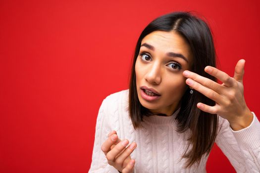 Closeup portrait of young beautiful brunette woman with short haircut with sincere emotions wearing casual sweater isolated over red background with copy space and touching eye.