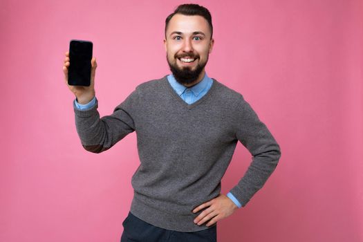 Handsome happy cool young man good looking wearing casual stylish clothes standing isolated over colourful background wall holding smartphone and showing phone with empty screen display looking at camera.