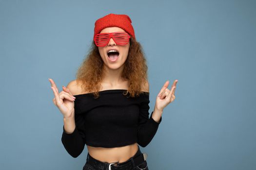 Shot of beautiful happy young dark blonde curly woman isolated over blue background wall wearing casual black crop top red bandanna and stylish red sunglasses looking at camera and having fun. copy space