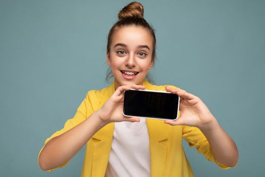 Photo of beautiful smiling girl good looking wearing casual stylish outfit standing isolated on background with copy space holding smartphone showing phone in hand with empty screen display for mockup looking at camera.