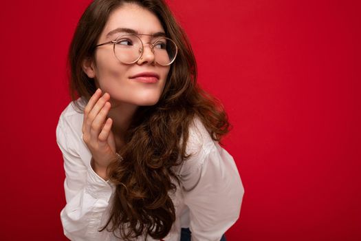 Photo shot of beautiful sexy young brunette curly woman isolated over red background wearing casual white shirt and stylish optical glasses looking to the side. copy space