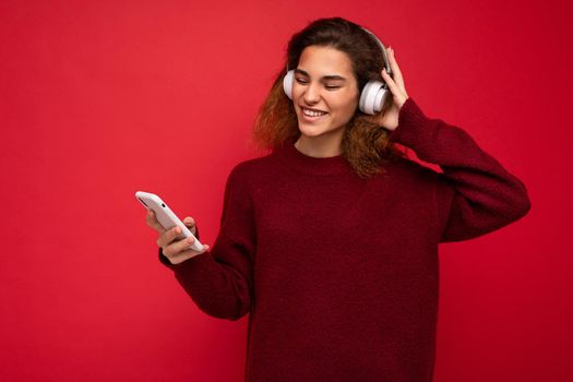 Shot of beautiful emotional young brunette curly woman wearing dark red sweater isolated over red background wall wearing white wireless headphones and listening to music and using mobile phone surfing on the internet and having fun.