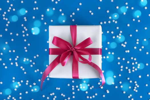 Gift box wrapped in white paper with a purple bow on festive blue background with many snowflakes. Copyspace for your text. Flat lay style. Christmas, New Year or birthday celebration concept