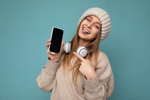 Photo of sexy attractive positive joyful young dark blonde woman wearing beige stylish sweater and knitted beige winter hat isolated on blue background wearing white bluetooth wireless earphones and listening to cool music and showing mobile phone with empty display for mockup looking at camera and pointing finger at smartphone.