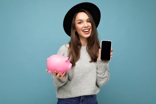 Portrait photo of happy positive smiling satisfied sincere young attractive brunette woman wearing stylish grey sweater and black hat isolated on blue background with free space and holding pink pig penny bank and showing mobile phone with empty screen display.