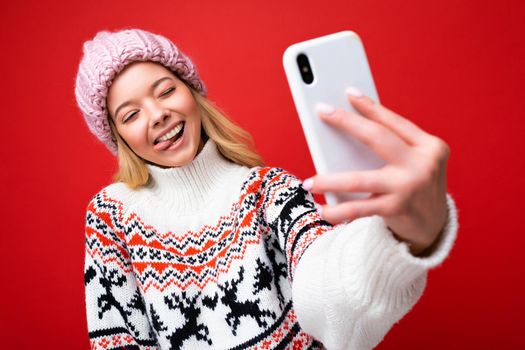 Photo of pretty positive young blonde woman wearing warm knitted hat and winter warm sweater standing isolated over red background using mobile phone taking selfie photo looking at device display and showing tongue.