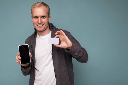 Handsome smiling blonde man wearing grey sweater and white t-shirt isolated over blue background wall holding credit card and mobile phone with empty screen for mock up looking at camera.