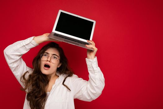 Photo shot of amazing funny happy beautiful smiling dark hair young woman holding computer laptop with empty monitor screen with mock up and copy space wearing white shirt looking up isolated over red wall background.