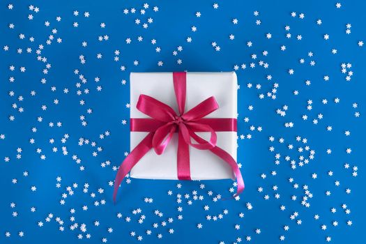 Gift box wrapped in white paper with a purple bow on festive blue background with many snowflakes. Copyspace for your text. Flat lay style. Christmas, New Year or birthday celebration concept