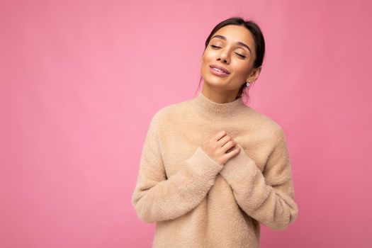 Portrait of young positive happy beautiful brunette woman with sincere emotions wearing casual beige jersey isolated over pink background with free space and holding hands on chest.